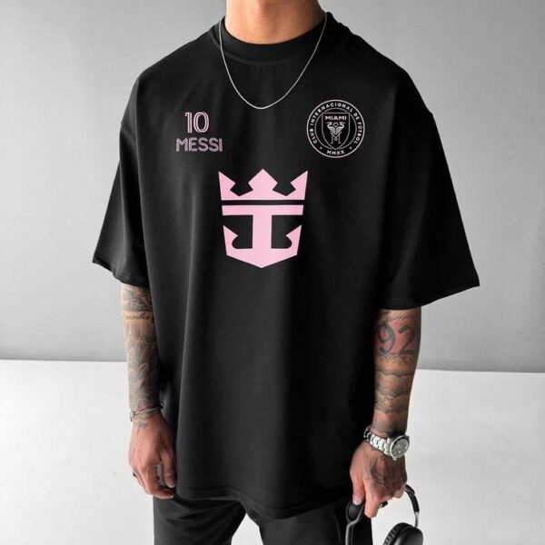 Oversized black T-shirt featuring a pink crown logo center, '10 MESSI' text on the left chest, and a circular emblem on the right chest; man wearing a chain and watch, holding headphones.