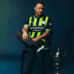 Man in a neon yellow-and-navy Manchester City jersey sits on a chair beside a silver trophy on the floor