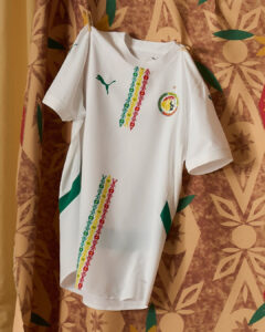 White soccer jersey hanging against patterned background, featuring a green Puma logo and circular crest on the chest with colorful diagonal stripe patterns down the front and side panels.