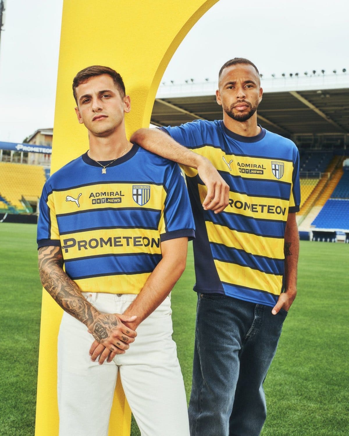 Two male soccer players in blue and yellow jerseys pose on a green field beside a large yellow arch in a stadium.