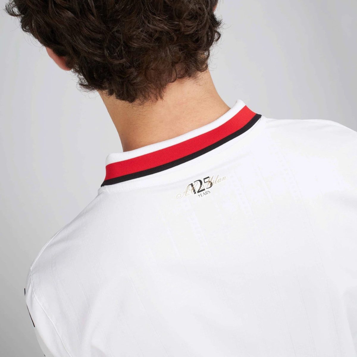 Back of a person wearing a white shirt with a red and black striped collar. The head is turned away, showing short curly hair at the nape.
