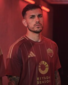Portrait of a man with short dark hair and a beard, wearing a maroon striped soccer jersey under red neon lights.