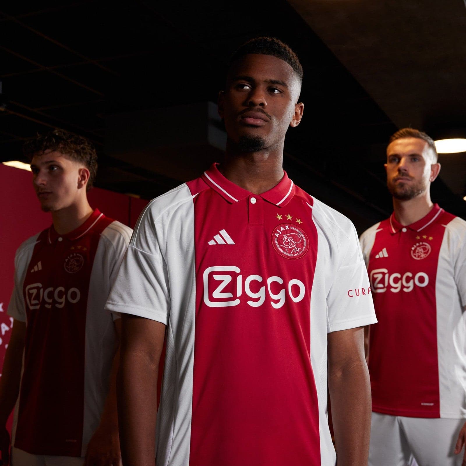 XfYZXKMBvxfTWe6 | UAE SPORT STORE Three soccer players in red-and-white Ajax jerseys stand in a dim hallway, facing forward.