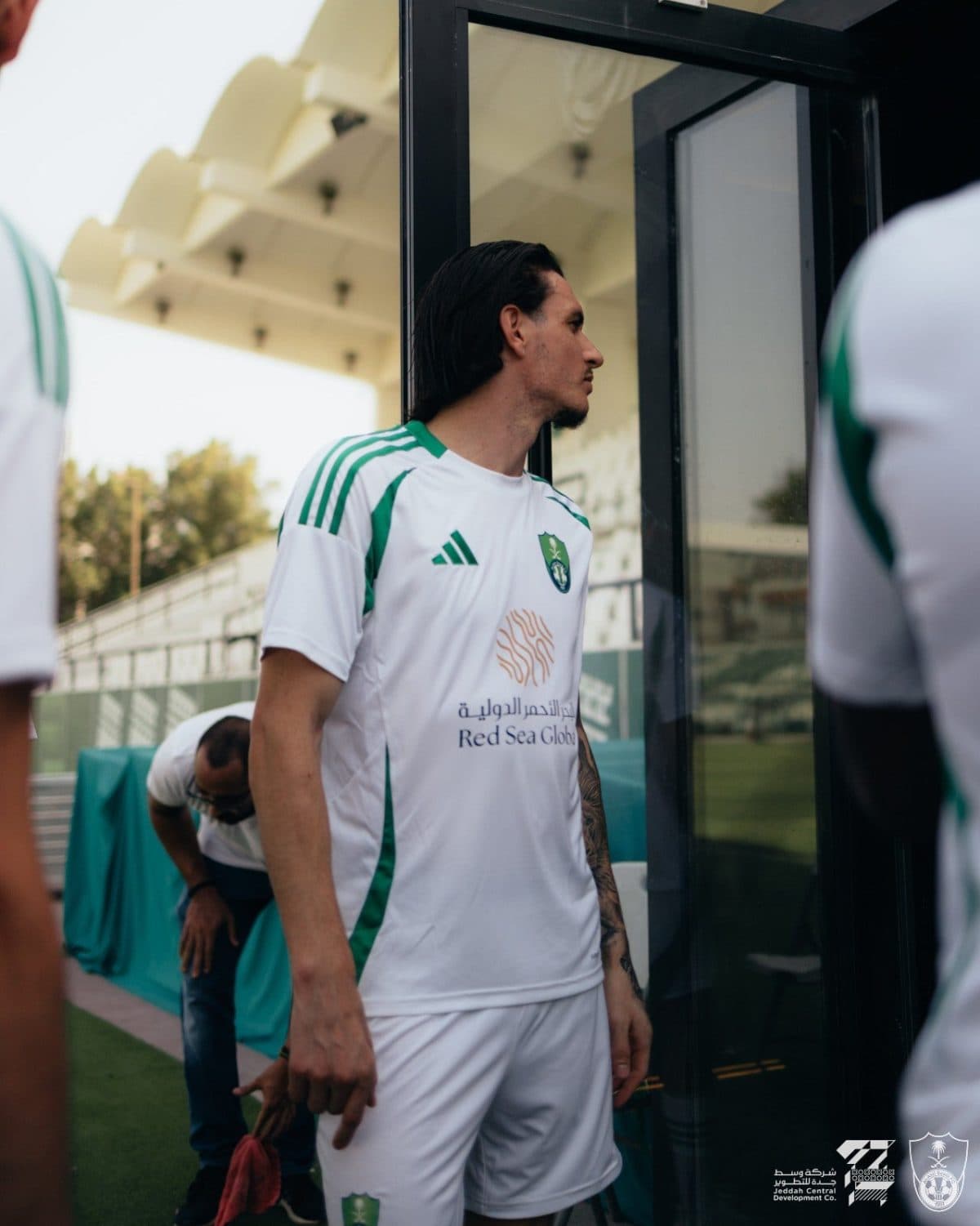 Athlete in white and green soccer kit standing beside a glass door, looking to the right, at a sports facility.