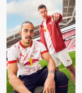 Two RB Leipzig players pose on a football pitch in a stadium; one seated in front, the other standing behind and pointing at the camera.