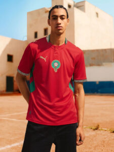 Portrait of a young man standing on a clay outdoor court, wearing a red soccer jersey with a crest and black shorts.
