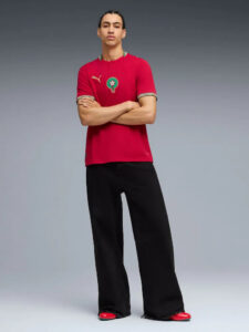 Young man stands with arms crossed, wearing a red short-sleeve shirt with a green-and-white team crest, black wide-leg pants, and red shoes against a gray gradient background.