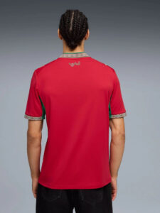 Back view of a person wearing a red polo with decorative patterned collar and sleeve cuffs, green side panels, and Arabic text on the upper back.