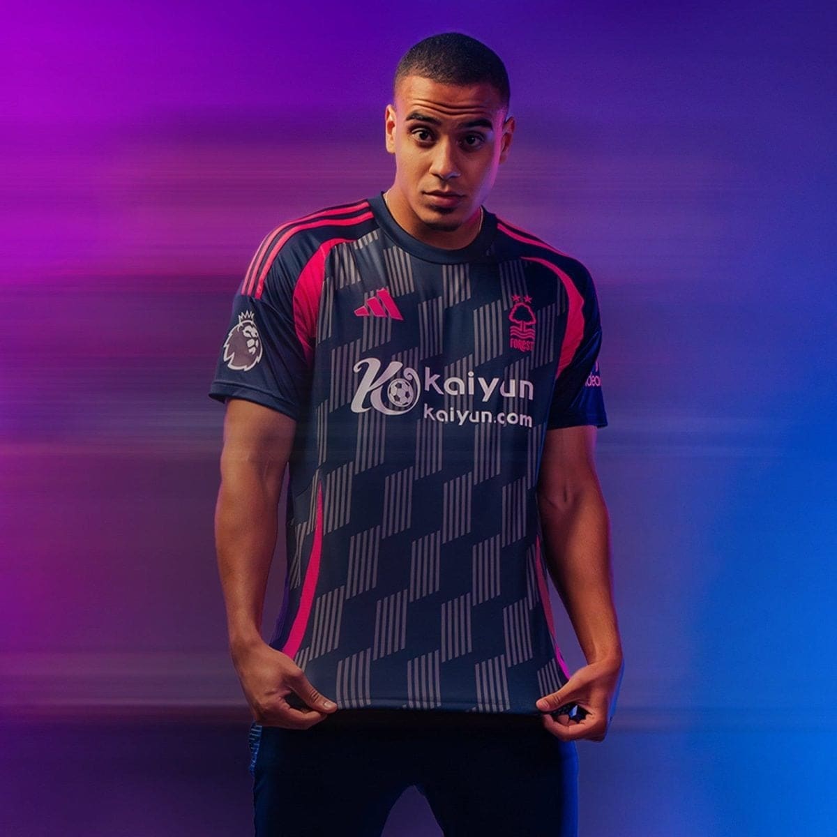 Athlete wearing a dark blue and pink soccer jersey with Kaiyun logo, standing with hands at his waist against a purple-pink motion-blur background