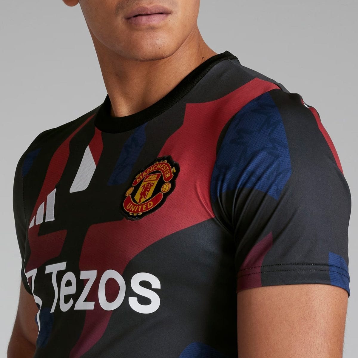 Close-up of a Manchester United football jersey with red and black diagonal stripes, showing the club crest and the Tezos sponsor logo on the chest area.