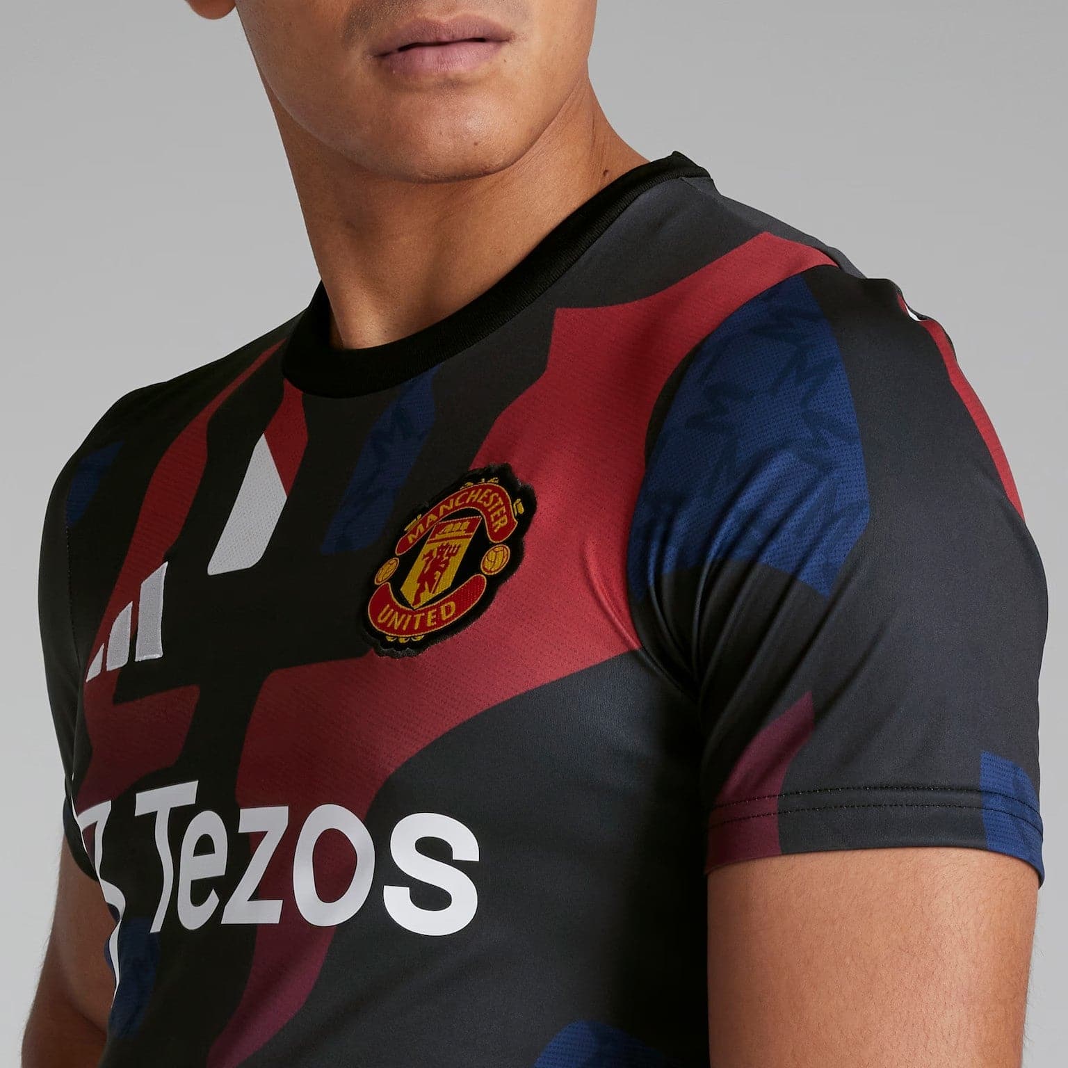 YFlKtRonDhEhvyS | UAE SPORT STORE Close-up of a Manchester United football jersey with red and black diagonal stripes, showing the club crest and the Tezos sponsor logo on the chest area.