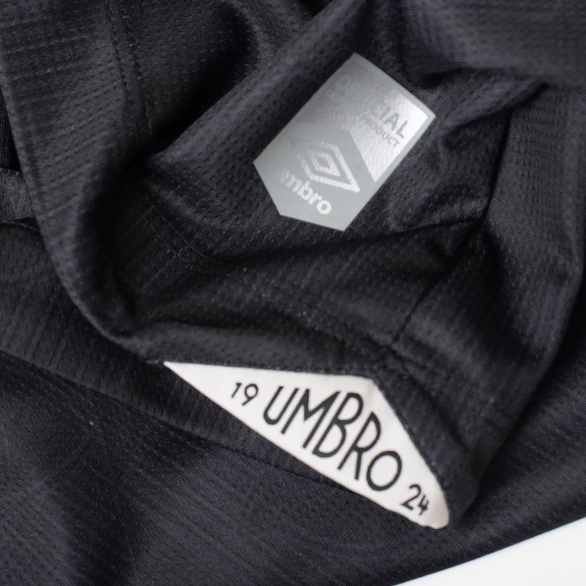 YHLZJV8AcrdW0lI | UAE SPORT STORE Close-up of a black garment's interior showing a white 'UMBRO' size tag with numbers 19 and 24.