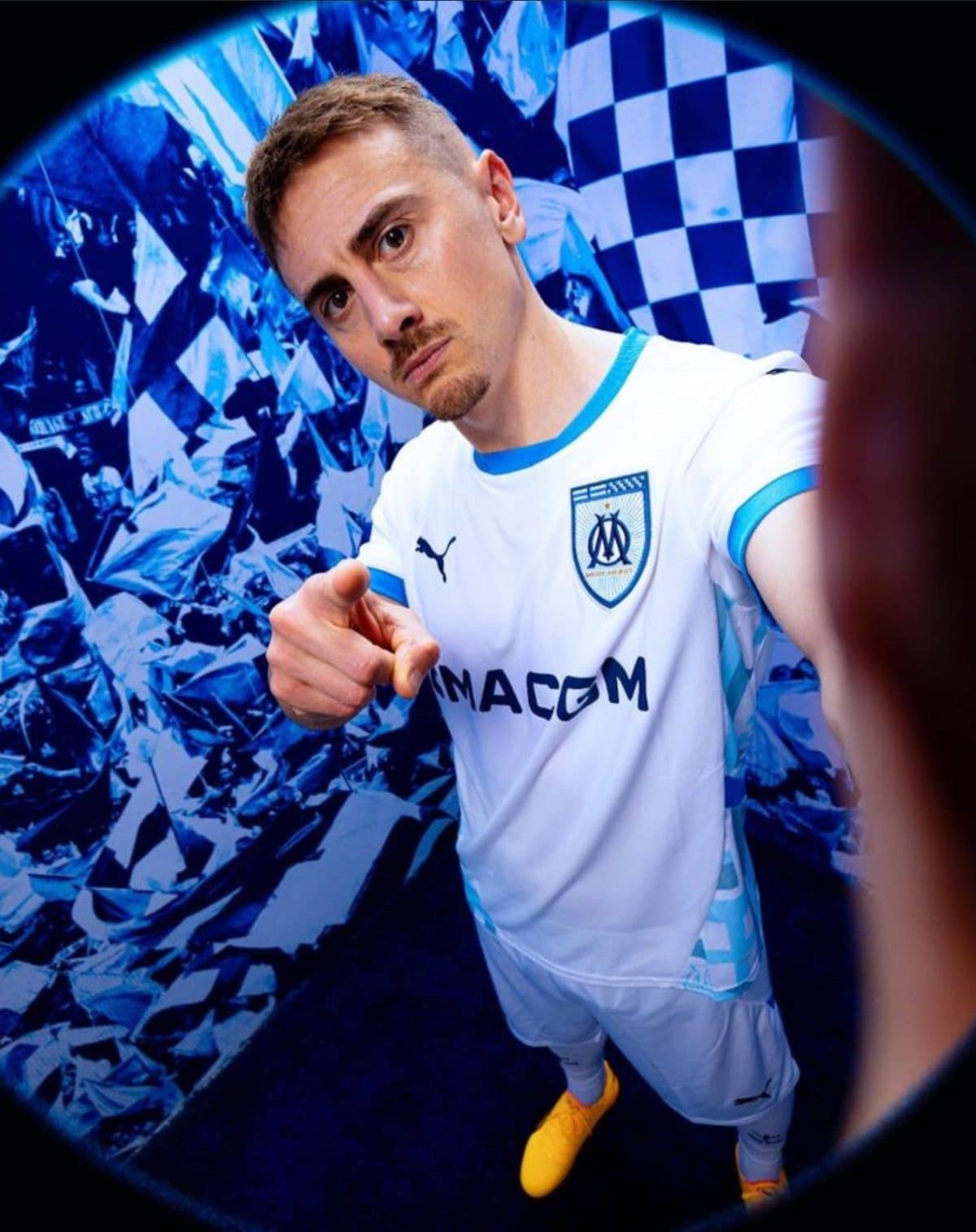 Footballer in a white Olympique de Marseille kit points at the camera against a blue metallic backdrop.
