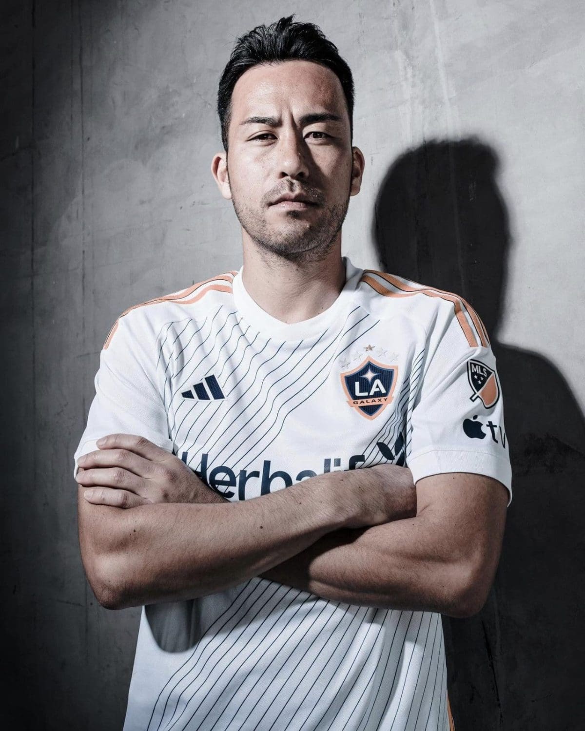 Portrait of a man in a white LA Galaxy soccer jersey with blue pinstripes, arms crossed, standing against a concrete wall.