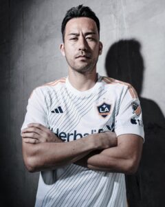 YJWV7gefU8PfhUd | UAE SPORT STORE Portrait of a man in a white LA Galaxy soccer jersey with blue pinstripes, arms crossed, standing against a concrete wall.