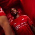 Close-up of a male footballer in a red jersey looking upwards in a tunnel or locker room, with team branding visible.