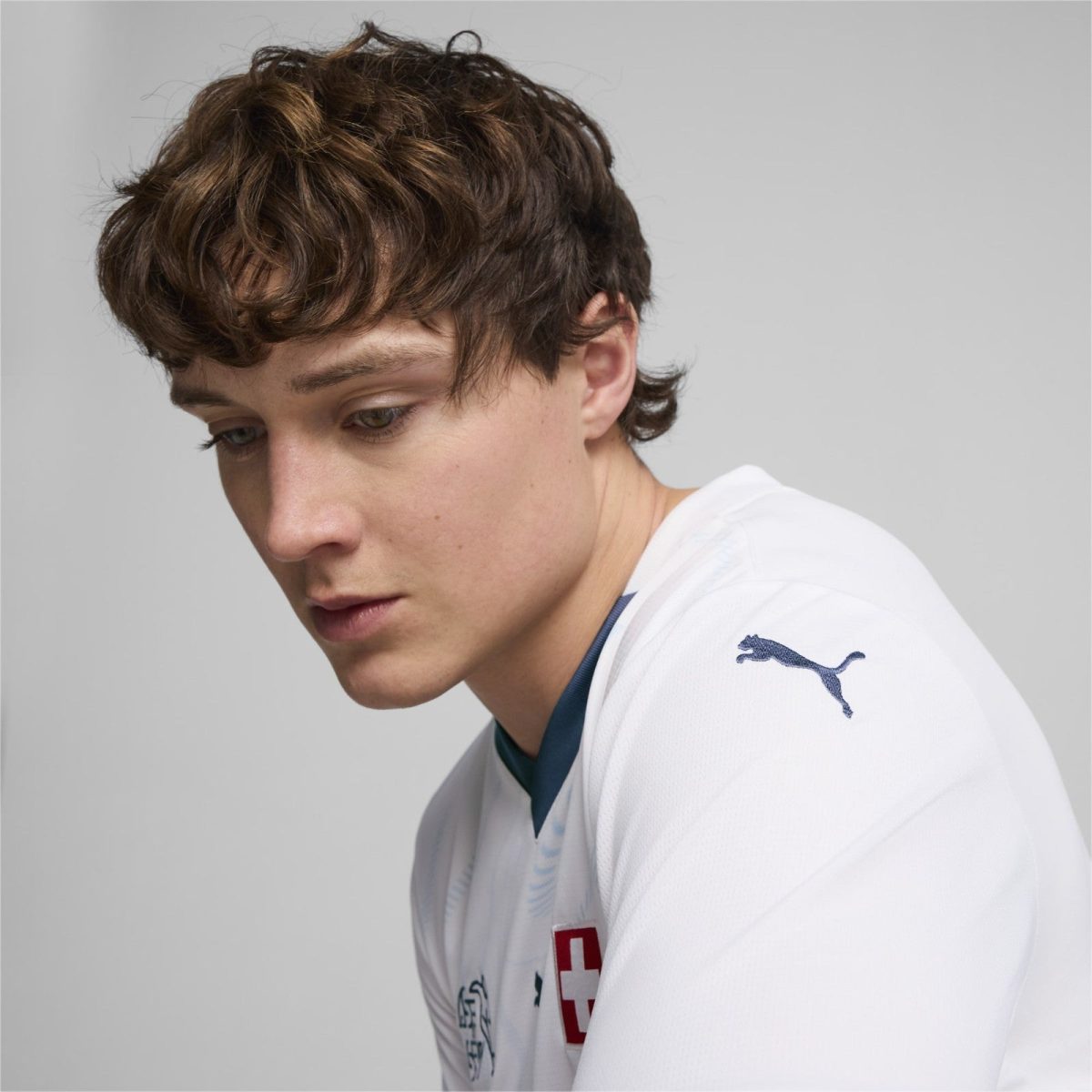 Young man with tousled brown hair wearing a white sports jersey, looking downward, Swiss emblem visible on chest and a blue collar detail.