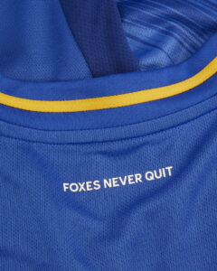Close-up of a blue sports jersey with yellow piping and the slogan 'FOXES NEVER QUIT' printed on it.