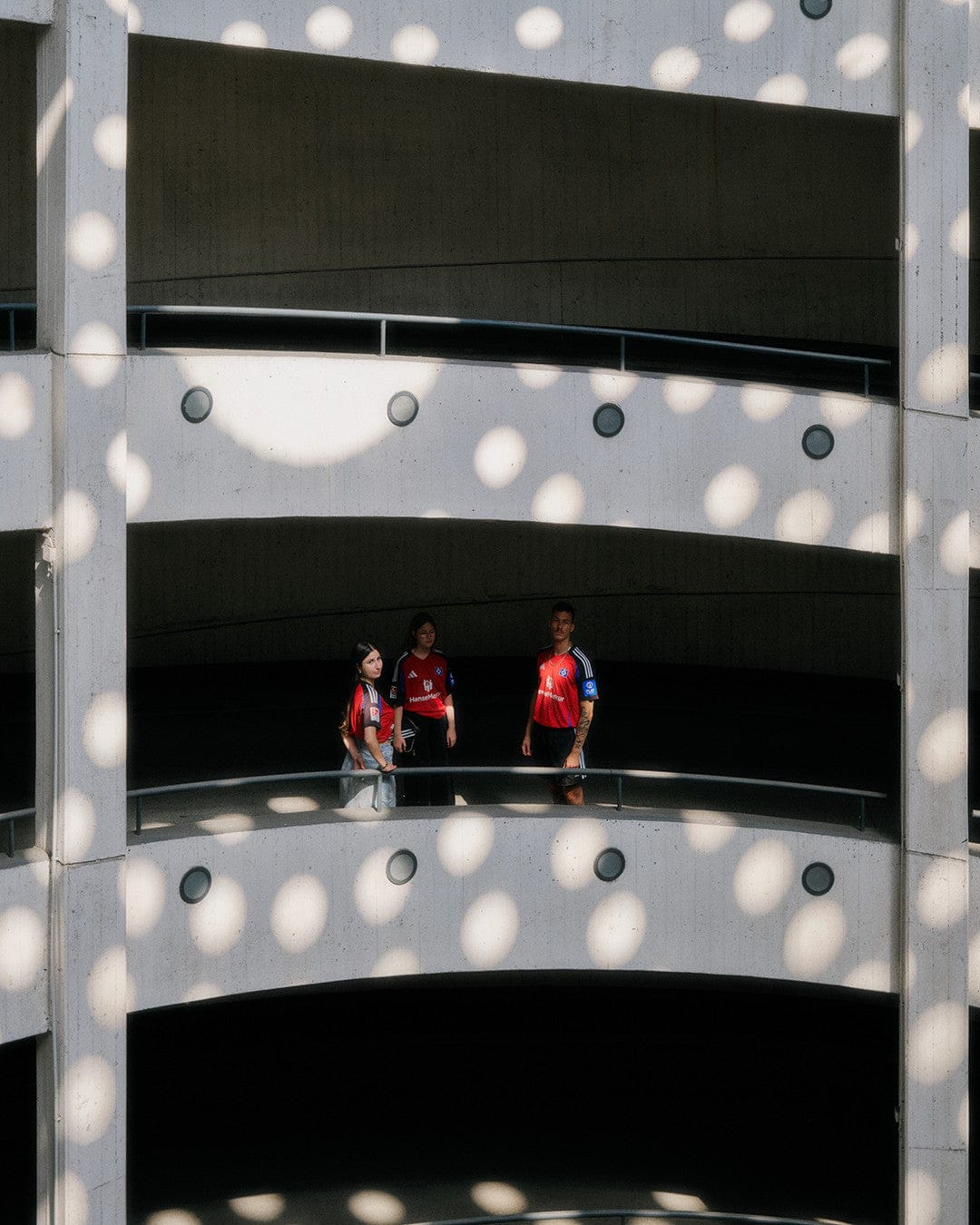 Z9TnwjWkVgYazaT | UAE SPORT STORE Three people in red jerseys stand on a curved concrete parking-garage balcony, bathed in circular light spots.