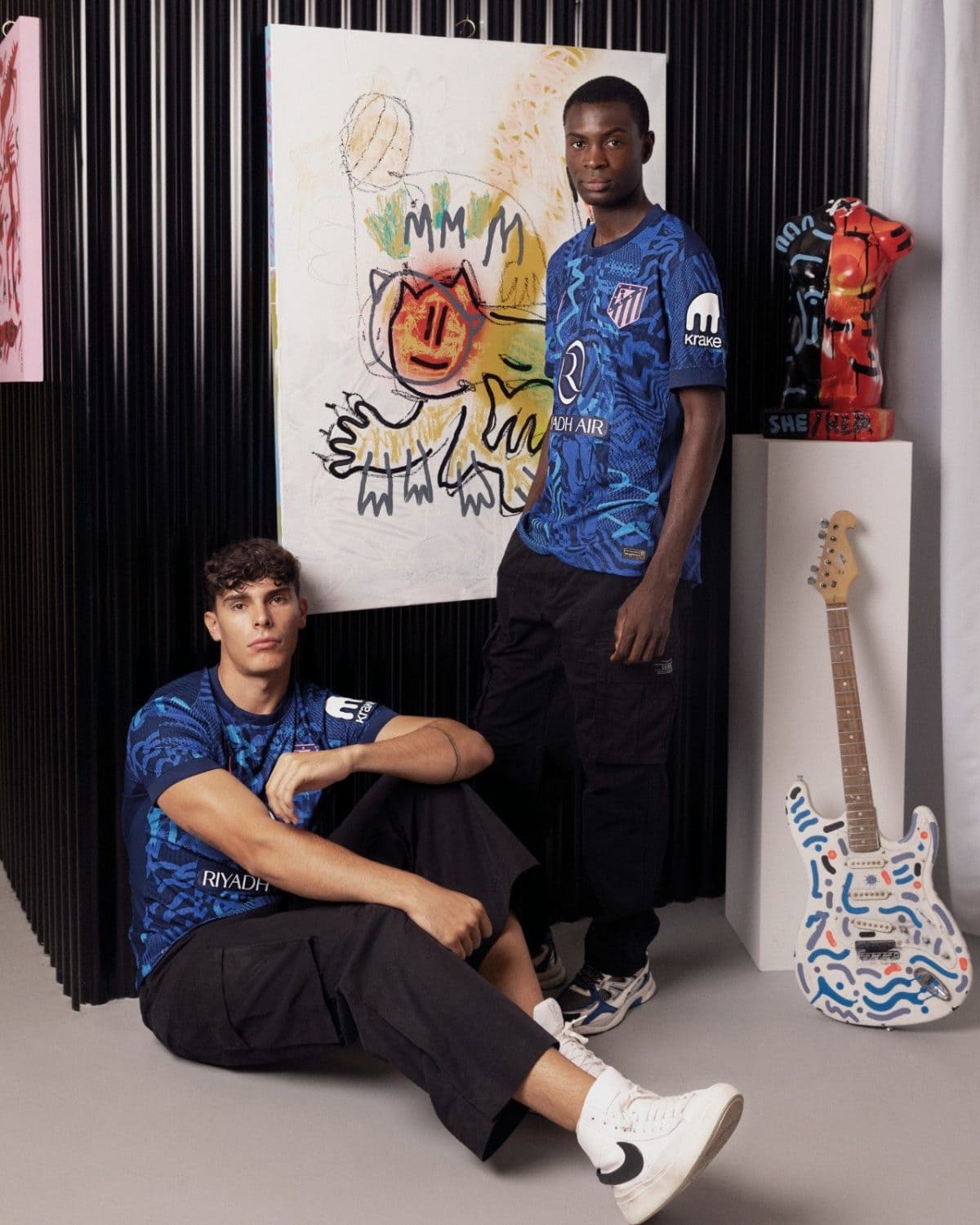 Two young men in blue Riyadh Air jerseys pose in a studio; a large abstract painting and a painted guitar on a pedestal nearby.