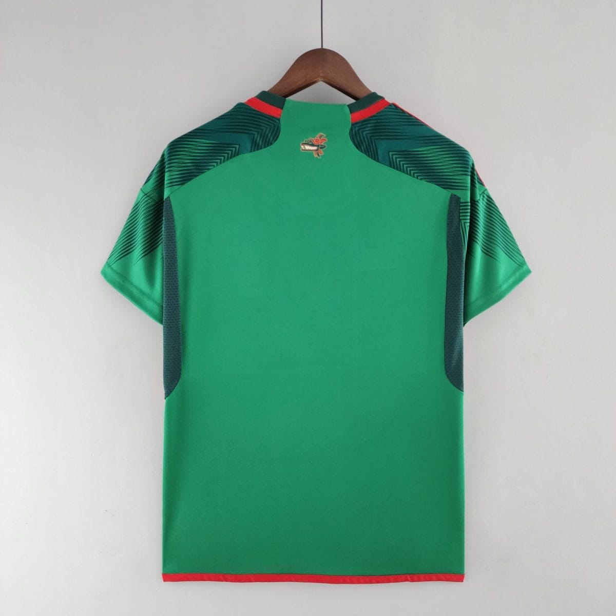 Back view of a green garment on a wooden hanger, with red trim at the hem and a small dragon emblem near the collar/back area, resting against a light wall.