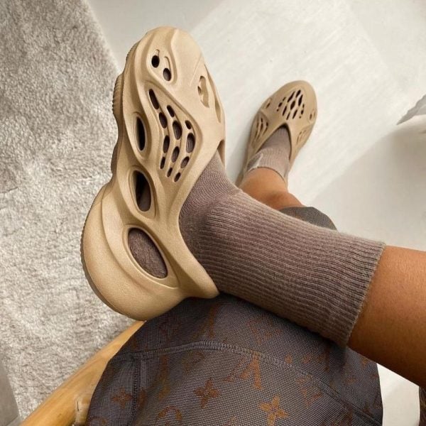 Beige slip-on sandals with cutouts resting on a crossed leg, wearing a brown sock and patterned pants nearby.