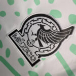 Embroidered circular patch with a pig and wing motif, black-and-white design, and the word MEXICO along a banner on the left.