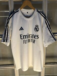 White Real Madrid soccer jersey on a hanger with Adidas logo, club crest, and 'Emirates Fly Better' sponsor text visible.