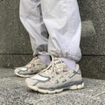 Person wearing light gray pants and white socks standing on a stone ledge, wearing beige/gray athletic sneakers with laces.