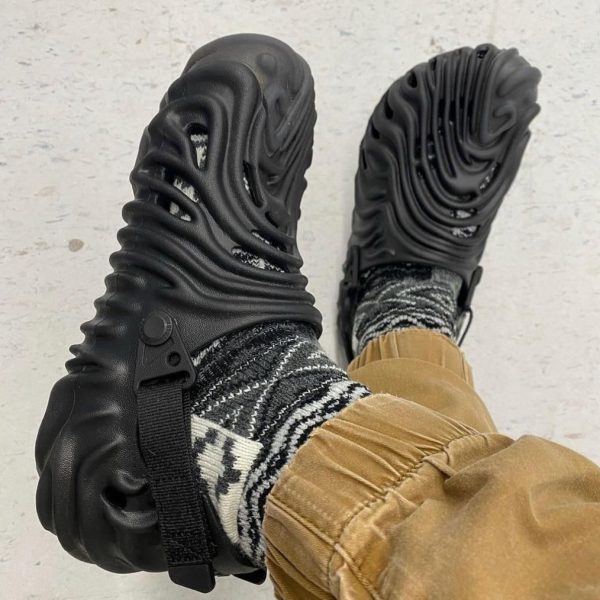 Black chunky sandals with cut-out, wave-like sole viewed from above, worn with patterned socks and tan pants.