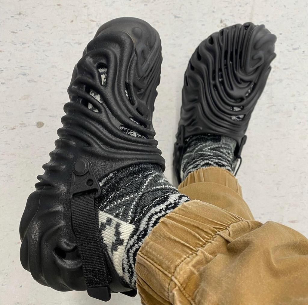 a10a4afe-1c1d-48a3-ba4f-ce0a901d6c9e | UAE SPORT STORE Black chunky sandals with cut-out, wave-like sole viewed from above, worn with patterned socks and tan pants.