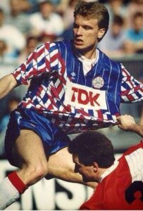 Soccer player in a blue and white checkered kit leaps over a defender in red, reaching for the ball with the crowd in the background.