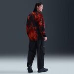 Person standing with back to camera, wearing a red and black patterned jacket, black cargo pants, and dark sneakers in a studio setting.