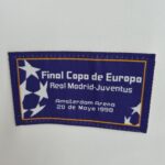 a2e0d9ab_2fe6c2cb-ef41-4698-aa68-dd3008c4f4a5 | UAE SPORT STORE Blue rectangular patch with a yellow border, white stars on the left, and white text reading 'Final Copa de Europa' and 'Real Madrid Juven...' with an Amsterdam reference (1998).