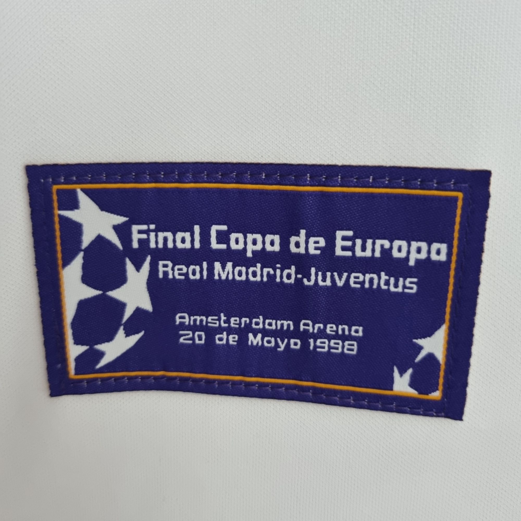 a2e0d9ab_2fe6c2cb-ef41-4698-aa68-dd3008c4f4a5 | UAE SPORT STORE Blue rectangular patch with a yellow border, white stars on the left, and white text reading 'Final Copa de Europa' and 'Real Madrid Juven...' with an Amsterdam reference (1998).