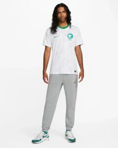 Full-body shot of a man wearing a white patterned soccer jersey with a green collar, gray joggers, and white-green sneakers.