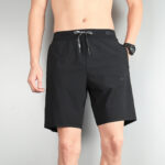 Man wearing black Nike athletic shorts with a drawstring waist, standing against a gray wall; watch on left wrist.