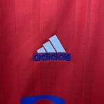 a40c3ebc_125568be-4253-4e45-baa7-29e66a2ab9bf | UAE SPORT STORE Close-up of red fabric featuring a partial blue Adidas logo with white stitching and stripes visible at the bottom.