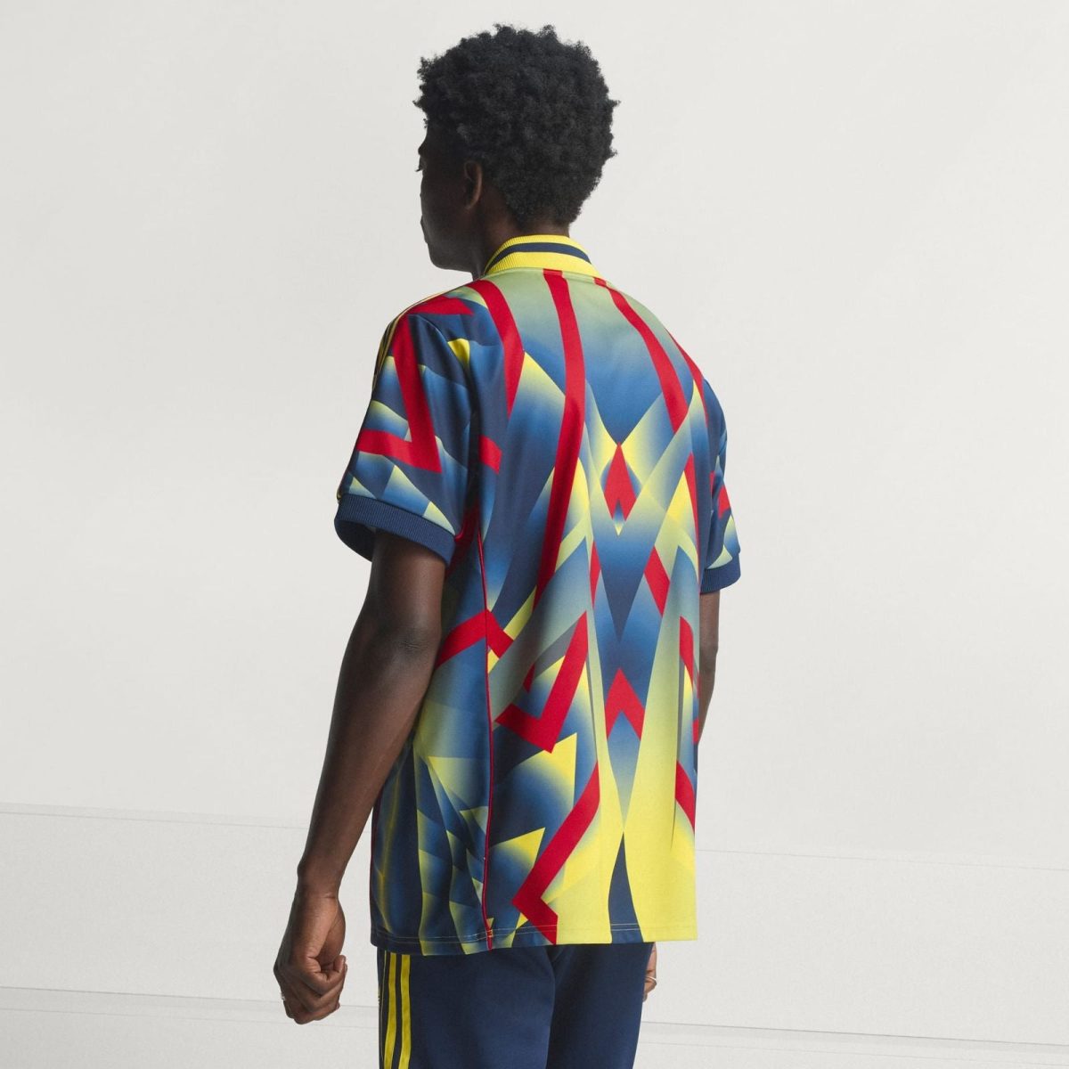 Person seen from behind wearing a bright geometric print shirt in blue, red, yellow, with a yellow collar trim against a neutral background.