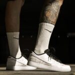 Close-up of a person wearing white Nike sneakers with black laces and white mid-calf socks, showing a tattooed leg.