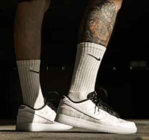 Close-up of a person wearing white Nike sneakers with black laces and white mid-calf socks, showing a tattooed leg.