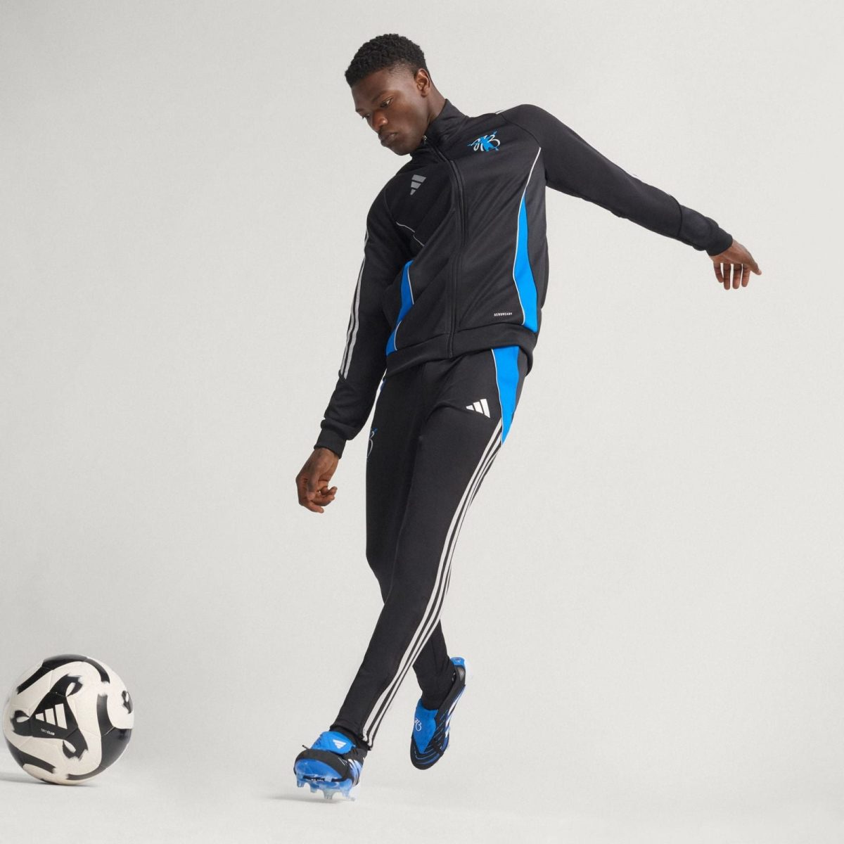 Male soccer player in a black tracksuit with blue accents kicking a soccer ball on a neutral background