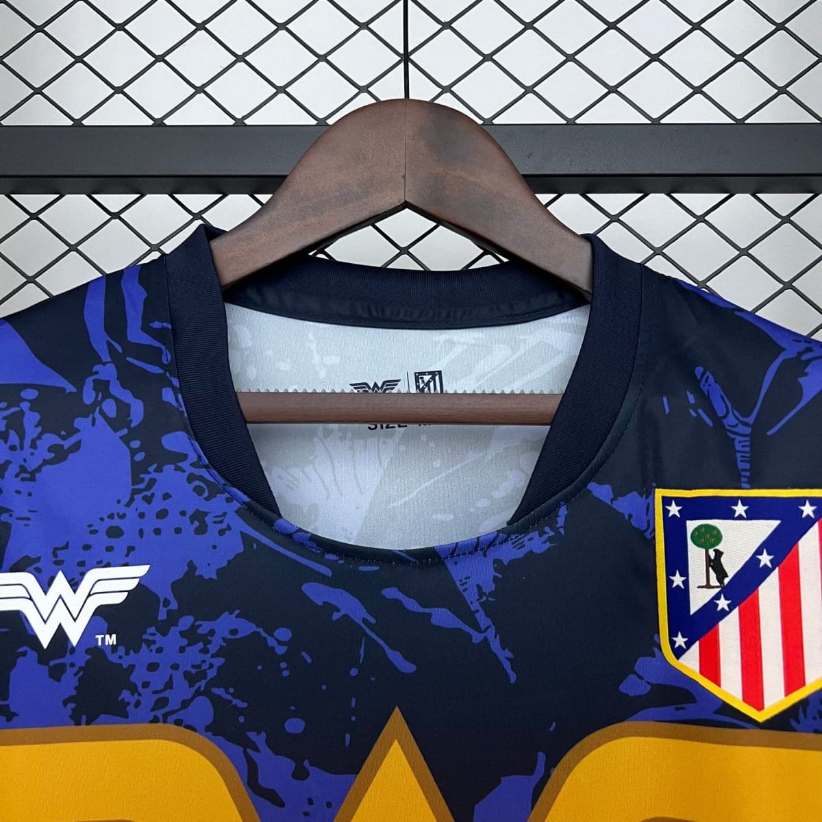 Navy blue athletic jersey with a purple abstract pattern on a wooden hanger, with a crest patch visible on the right and a chain-link fence behind.