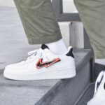 White Nike Air Force 1 sneaker with an orange overlay on the swoosh, worn with white socks and olive-green pants stepping onto a stone ledge.