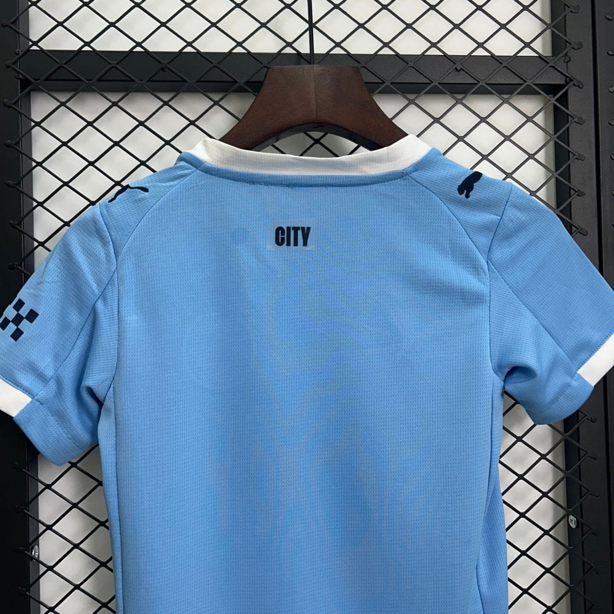 Light blue athletic jersey on a wooden hanger, displaying a 'CITY' label, with dark brand marks on the shoulders, hanging in front of a metal chain-link fence.