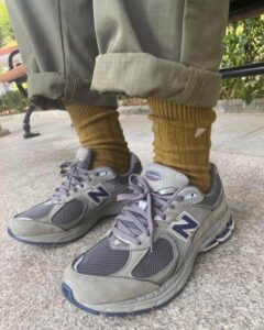 a61f1e97-fbc5-4a4b-9652-4a09227c3f55 | UAE SPORT STORE Close-up of gray New Balance sneakers with navy N logos, mustard socks, and rolled khaki pants outdoors on a concrete surface.