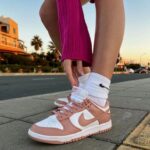 Close-up of white and pink Nike sneakers on a person’s feet, standing on a sidewalk with a city street in the background at sunset.
