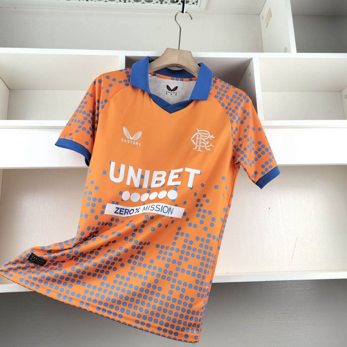 Bright orange sports jersey with blue polka dots hangs on a wooden hanger in a white closet; features a blue collar and sponsor logos including UNIBET and a club crest on the chest.