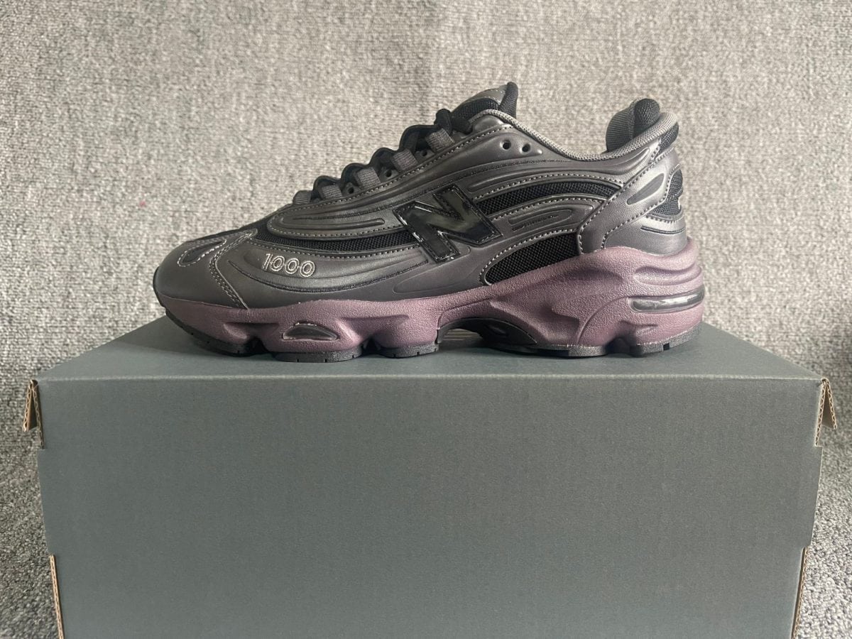 Black chunky athletic sneaker with gray stitching and mesh panels resting on a dark gray shoebox against a textured gray backdrop.