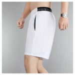 a6c96f29 | UAE SPORT STORE Male model wearing white athletic shorts with a black elastic waistband (Under Armour) and a side pocket visible on the left hip, knee-length cuffed hem.
